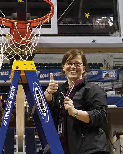 Noel Devin on a ladder underneath a basketball hoop giving thumbs up